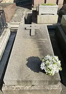Photo d'une tombe comportant une croix en relief sur la partie supérieure, une liste de noms gravés en doré au centre ainsi qu’un bouquet de fleurs en bas à droite