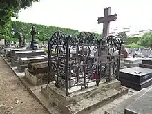 Photographie d'une tombe entourée d'une grille en fer et surmontée d'une croix.