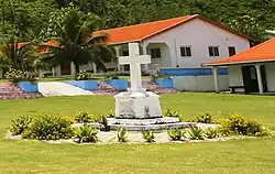Photo en couleur montrant une tombe blanche placée sur une pelouse et entourée de petits buissons et surmontée d'une croix blanche ; en fond, des maisons et des palmiers.