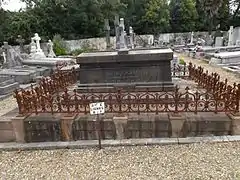 Photographie d’un tombeau entouré d’une grille dans un cimetière.