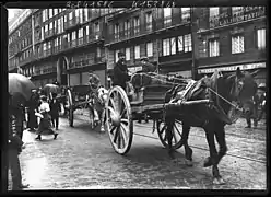 File de charrettes tirés par des chevaux remontant une rue.