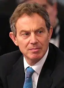 Photographie d'un homme grisonnant, il porte une veste noire, une chemise blanche et une cravate bleue.