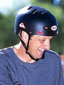 Tony Hawk souriant, en gros plan avec un t-shirt gris, un casque de skateboard, pris de face avec la tête tournée vers sa gauche, légèrement inclinée vers le bas. On distingue le soleil dans le cou du skater, ainsi que son reflet sur le casque.