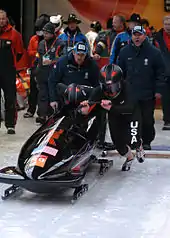 Photographie d'un bobsleigh aux couleurs américaines poussés par deux bobeurs, derrière lesquels se trouve plusieurs hommes.