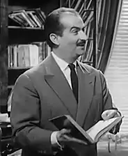 Photogramme d'un film en noir et blanc. Un homme âgé d'une quarantaine d'années, cheveux clairsemés et petite moustache, en costume cravate. Il tient un livre ouvert dans ses mains et sourit à quelqu'un hors cadre, à droite.