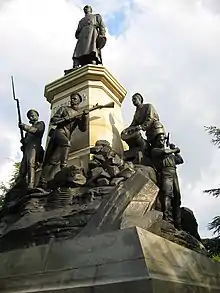 Photographie d'une statue en bronze d'un homme portant un long uniforme au sommet d'un pilier blanc. Au pied de cette dernière, des statues représentent des soldats manœuvrant un mortier, tenant un fusil ou utilisant une pioche.