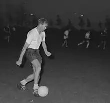 Photo en noir et blanc d'un joueur de football en maillot blanc et short bleu marine en action, conduisant le ballon de son pied droit.