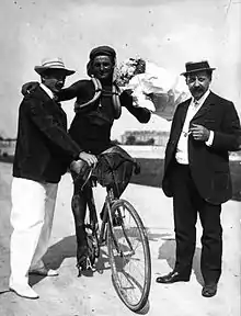 Photographie noir et blanc d'un cycliste à l'arrêt entouré de deux hommes debout.