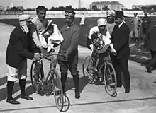 Photographie en noir et blanc de deux cyclistes tenant un bouquet de fleurs dans leurs mains, des hommes soutenant leur vélo.