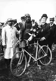 Photographie noir et blanc d'un coureur avec son vélo entouré d'officiels.