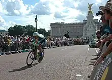 Photographie couleur d'un cycliste sur son vélo, en tenue de cycliste bleue et verte, salué par la foule dans une rue de Londres, sous un ciel bleu nuageux.