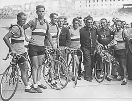 Photographie en noir et blanc d'un groupe de cyclistes se tenant debout à côté de leurs vélos à l'arrivée d'une course.