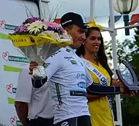 Photograhpie de trois-quarts d'un coureur cycliste, habillé en blanc, souriant, levant un bouquet de fleurs.