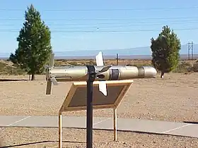 Photographie en couleurs d'un missile gris métallisé reposant sur un support noir devant une pancarte, deux arbres en arrière-plan