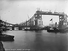 photographie en noir et blanc montrant un pont en construction, au premier plan des bateaux