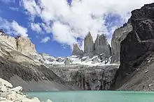 Un resserement dans la vallée bloque un glacier et préserve un grand lac de montagne. Au-dessus trois pics neigeux volent dans les nuages.