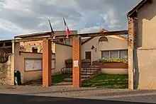 Photo de la mairie : deux colonnes en briques rouges devant les marches qui mènent à la porte du bâtiment. "Mairie" est inscrit sur le fronton, au-dessus des drapeaux français et européen.