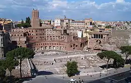 derrière des ruines au ras du sol se dresse une place semi-circulaire à colonnades sur deux étages, au-delà de laquelle s'étendent de plus hauts bâtiments, également en briques.