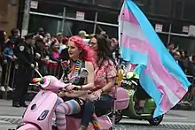 Deux personnes sur une mobylette avec un drapeau trans. Une des personnes porte des collants avec les couleurs trans (juin 2019).