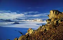 Vue plongeante depuis une hauteur rocheuse sur un paysage glaciaire parsemé de montagnes, sous un ciel bleu profond.