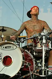 photo d'un homme torse nu tatoué portant une casquette rouge et jouant de la batterie.