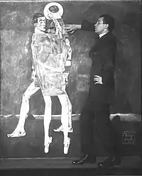 Photo noir et blanc d'un homme debout de profil, main gauche sur la hanche, la droite sur une toile au mur avec deux grands personnages