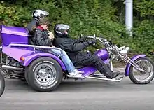 Photo couleur d'un tricycle motorisé, avec un conduxteur casqué et deux passagers.