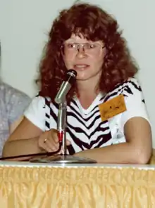 Photo couleur d'une femme blanche à lunettes et aux cheveux longs bouclés en gros plan, assise à une table.
