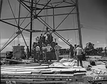 Photographie d'un groupe d'hommes entourant une sphère accrochée à un câble à la base d'une tour métallique.