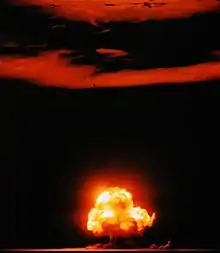 Explosion au niveau du sol et début de la formation d'un champignon atomique. Les nuages et le sol sont illuminés par la couleur orange de l’explosion.
