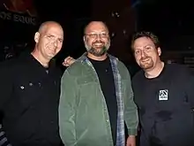 Trois hommes souriants photographiés la nuit, dont deux sont habillés en noir et le dernier en vert.