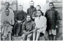 Photo en noir et blanc montrant trois prêtres français entourés de cinq séminaristes wallisiens, assis sur des marches et prenant la pose.