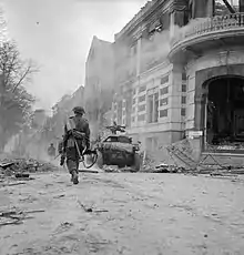 Un soldat marche en s'éloignant de la caméra, au long d'une rue jonchée de with debris. Plus loin, un petit véhicule blindé.