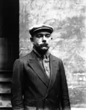 Portrait d'un homme vêtu d'un costume et d'une casquette, se tenant debout.