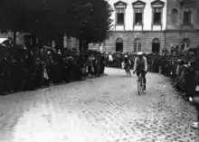 Photographie en noir et blanc montrant deux cyclistes traversant une ville sur une rue pavée, au milieu de la foule.