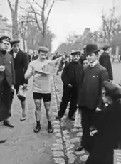 Photographie en noir et blanc d'un cycliste lisant un journal, entouré d'autres hommes.