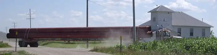 Un camion franchit une intersection dans une zone rurale avec, entreposés sur sa remorque, quelques tuyaux d'une douzaine de mètres de longueur.