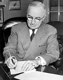 Un homme en costume gris et portant des lunettes signe un document