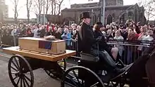 Une calèche passe devant des badauds. L'arrière est constitué d'une plate-forme ouverte sur laquelle est posé un cercueil recouvert d'un drapeau.