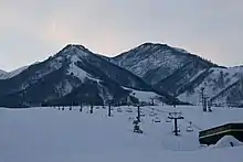 Photo couleur de deux montagnes sous un ciel blanc laiteux avec des pentes skiables et une remontée mécanique au premier plan.