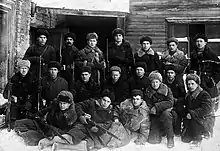 Photo noir et blanc d'une vingtaine d'hommes en chapka posant devant un bâtiment, certains tiennent un fusil