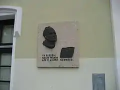 Photo couleur d'une plaque de marbre clair sur un mur, avec une tête en haut-relief et une inscription en noir