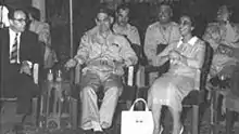 Deux hommes et une femme en costume, uniforme militaire, et robe, assis en cercle.