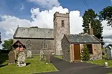 Photo d'une église comprenant une petite tour attachée à un bâtiment moins haut. Sur la gauche, se trouvent des pierres tombales alors que sur la droite se trouve un petit bâtiment, situé devant l'église, avec une porte marron.