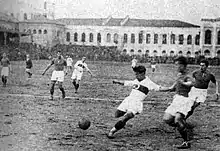 Photographie en noir et blanc d'une scène d'un match de football. Un joueur s'apprête à tirer.