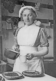 Photographie monochrome de Turner portant un tablier de cuisine et un chapeau. Elle tient une casserole à sauce et un verre à mesurer. Sur la table devant elle sont disposés plusieurs flancs dans des plats en métal.