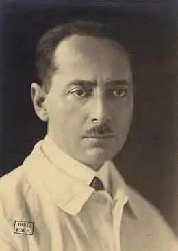 Photographie en noir et blanc d'un homme portant blouse blanche par-dessus son costume, et arborant un regard soucieux.