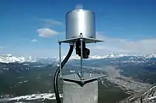 Un cylindre métallique repose sur une plate-forme carrée soulevée par deux boulons. Un prisme relié à des câbles surveille les mouvements de la montagne.