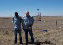 Deux hommes, l'un Noir et l'autre Blanc, se recueillant sur les lieux de la catastrophe, une prairie sèche et ensoleillée. Une ancienne structure de la mine se trouve derrière eux.