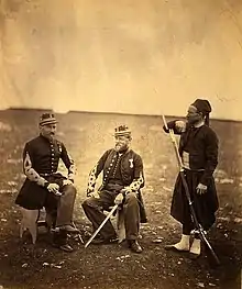 Photographie de deux officiers assis sur des tabourets en bois et d'un zouave reconnaissable à son turban, son pantalon bouffant et ses guêtres.